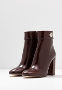 Botas de tobillo de piel marrón con un acabado brillante, punta afilada y tacón de madera apilada. Presenta un detalle de botón metálico en la parte superior.