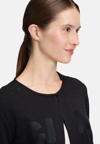 Kobieta z brązowymi włosami związanymi z tyłu nosi czarny kardigan z małymi czarnymi koralikami oraz dużymi literami "G" i "A" z przodu.