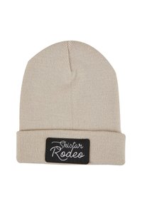 Krémová pletená čepice s ohrnutým lemem. Obsahuje obdélníkovou černou nášivku s bílým textem "Skistar Rodeo." Měkká textura a teplý design.