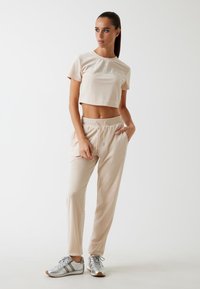 Vaaleanbeige crop-top, jossa on teksturoitu logo; matchaavat beige collegehousut, joissa on joustava vyötärö ja nyöri; hopeiset urheilukengät.