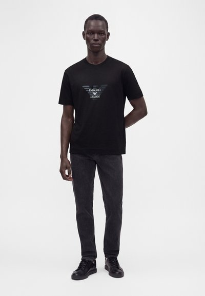 Άνδρας που φοράει μαύρο T-shirt με το λογότυπο Emporio Armani, σκούρα τζιν και μαύρα αθλητικά παπούτσια, στέκεται μπροστά σε ένα απλό φωτεινό φόντο.