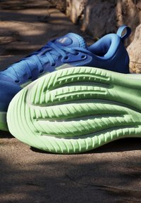 Chaussure de course bleue avec une semelle texturée vert clair, posée sur le sol extérieur près des racines d’arbre sous la lumière du soleil.