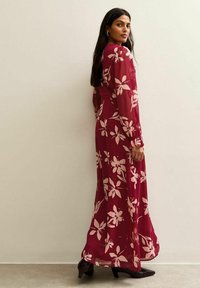 Vrouw in een lange bordeauxrode jurk met lichte bloemenprint, zwarte puntige hakken, staande tegen een effen beige muur en over haar schouder kijkend.