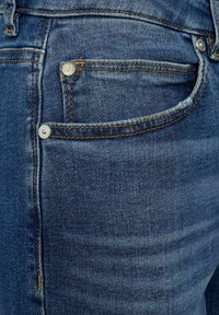 Jeans de mezclilla azul oscuro con una textura suave, costuras de cobre y acentos de hardware plateado en los bolsillos y el cierre de botón.