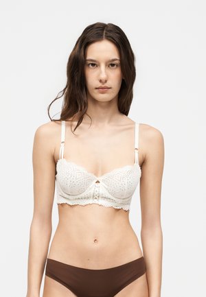 Kvinde med langt brunt hår iført en hvid blondetop og brune undertøj, stående mod en ensfarvet lys baggrund.