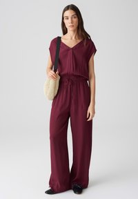 Burgunderfarbener Einteiler mit kurzen Ärmeln, V-Ausschnitt, elastischem Bund und weitem Bein. Das Model hält eine runde, geflochtene Tasche mit einem Trageriemen. Schwarze Schuhe.