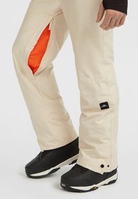 Beige skibroek met een oranje mesh voering, voorzien van ritsen en een logo-patch. Zwarte sneeuwlaarzen met gestructureerde zolen zijn ook zichtbaar.
