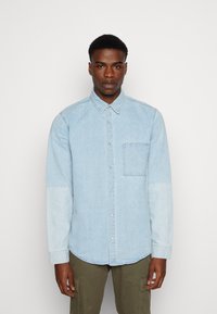 Camicia in denim azzurro chiaro con colletto a punta, taschino sul petto e sezioni delle maniche di un tono più chiaro. Texture liscia, maniche lunghe, orlo curvo.