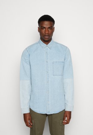 Camicia in denim azzurro chiaro con colletto a punta, taschino sul petto e sezioni delle maniche di un tono più chiaro. Texture liscia, maniche lunghe, orlo curvo.