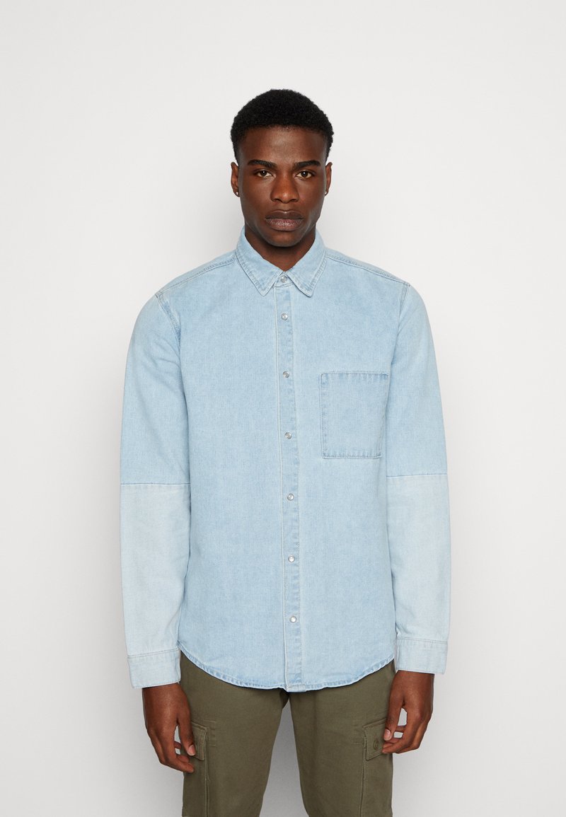 Camicia in denim azzurro chiaro con colletto a punta, taschino sul petto e sezioni delle maniche di un tono più chiaro. Texture liscia, maniche lunghe, orlo curvo.