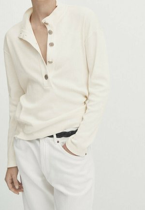 Une personne portant une chemise henley à manches longues de couleur crème, avec trois boutons déboutonnés, et un pantalon blanc avec une ceinture tressée noire.