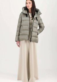 Donsjack in licht olijfgroen met een capuchon, voorzien van een gestikte ontwerp, vooraan met een ritssluiting en verstelbare koordjes. Gecombineerd met beige wijde broek.