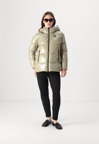 Casaco verde-azeitona puff e brilhante, com capuz, frente com fecho éclair e dois bolsos laterais, vestido sobre calças pretas e sapatos pretos.