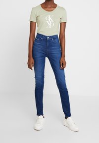 Grön t-shirt med vitt logotyp, högmidjade blå skinny jeans och vita sneakers. Slätt tyg; åtsittande design; avslappnad look.