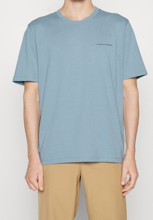 Homme portant un t-shirt bleu clair à manches courtes avec le texte « Tiger of Sweden » sur le côté gauche de la poitrine et un pantalon beige.