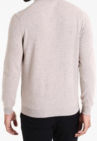 En person som bär en ljusbeige långärmad tröja och svarta byxor, sedd bakifrån.