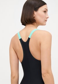 Μαύρο μαγιό με σχέδιο racerback, που διαθέτει τιράντες σε τουρκουάζ απόχρωση και αντίθετη ραφή. Το υλικό φαίνεται λείο με εφαρμοστό σχήμα.