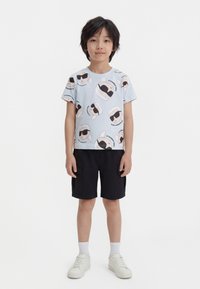 Băiat stând în picioare, purtând un tricou bleu cu fețe de desene animate, pantaloni scurți negri cu buzunare, șosete albe și adidași albi, pe un fundal alb.