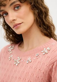 Pull en rose avec un design en tricot torsadé texturé. Comprend des ornements décoratifs de gemmes et de perles le long de l'encolure.
