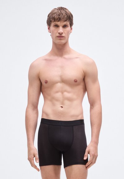 Cuecas boxer pretas com textura suave e cintura elástica, apresentando um design ajustado. O modelo exibe uma postura neutra.