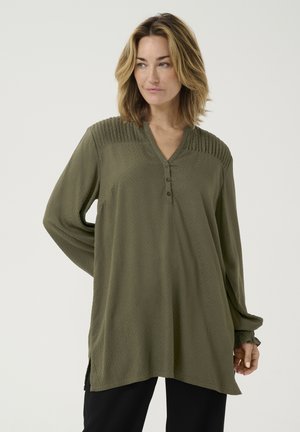 Tunique vert olive à boutons, avec un motif en losanges texturé, décolleté en V, manches longues et fentes latérales pour faciliter les mouvements.
