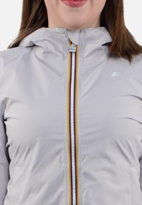 Giacca grigia chiara con zip frontale, dotata di cappuccio, cerniera a strisce multicolori e un piccolo logo sul petto. Texture liscia.