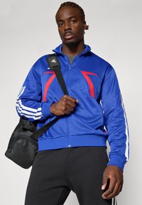 Blå zip-up treningsjakke har røde detaljer og hvite striper på ermene. Modellen holder en svart treningsbag med skulderrem.