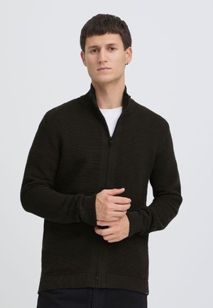 Jaunas vyras su trumpais tamsiais plaukais, dėvintis juodą ribuotą megztinį su užtrauktuku ant balto marškinio, stovintis priešais paprastą šviesią foną.