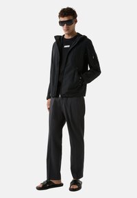 Czarna lekka kurtka z kapturem, z zamykanymi kieszeniami, połączona z czarną koszulką, ciemnoszarymi spodniami i czarnymi sandałami. Elegancki, minimalistyczny design.