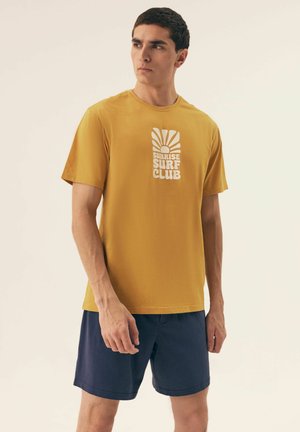 T-shirt z krótkim rękawem w kolorze musztardowym z białą grafiką "Sunrise Surf Club". W połączeniu z granatowymi szortami, gładkim materiałem i luźnym krojem.