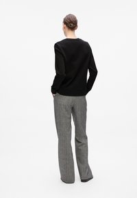 Une personne avec les cheveux en chignon se tient de dos, portant un haut noir à manches longues et un pantalon large gris, les mains dans les poches.