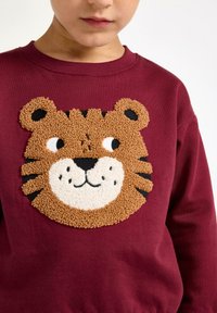 Bambino che indossa un maglione bordeaux con un'applicazione di un volto di tigre sorriso in rilievo sul petto, su uno sfondo bianco semplice.