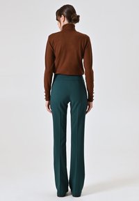 Maglione a collo alto di colore marrone scuro, dalla forma aderente. Abbinato a pantaloni a vita alta e svasati di colore verde profondo, in tessuto liscio, con dettagli minimi.