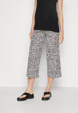 Vrouw met een zwarte mouwloze top, zwart-wit gevlekte cropped broek met wijde pijpen, en zwarte platform sandalen.