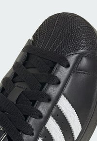 Čierna teniska s textúrovanou čiernou špičkou, čiernymi šnúrkami a bielymi pruhmi. Hladký kožený zvršok so záberom zblízka na detaily stehov.