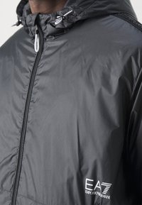 Chaqueta cortavientos negra Emporio Armani EA7 con cremallera y capucha, con toggles de cordón blancos, llevada en la parte superior del torso.