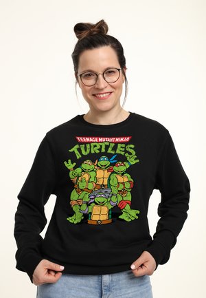Sort sweatshirt med et farverigt grafisk motiv af fire skildpadder fra "Teenage Mutant Ninja Turtles," med tekst ovenfor i rød og gul.