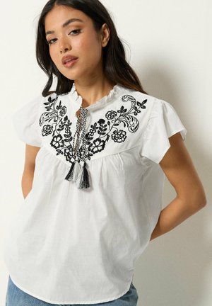 Woman wearing white short-sleeve blouse with black floral embroidery and tassel ties, posing against a plain light background.