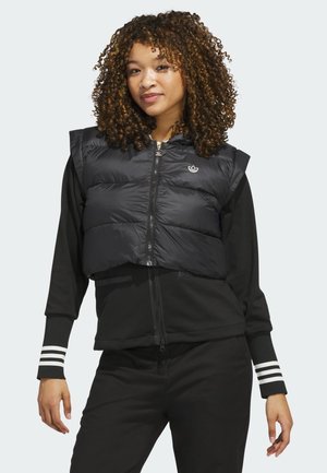 Joven con cabello rizado que lleva un chaleco negro corto acolchado sobre una chaqueta negra con cremallera y pantalones negros sobre un fondo claro.