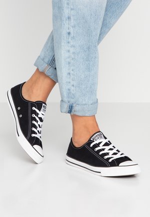 Person trägt hochgekrempelte hellblaue Jeans und schwarze Converse-Low-Top-Sneaker mit weißen Schnürsenkeln und steht auf weißem Boden.