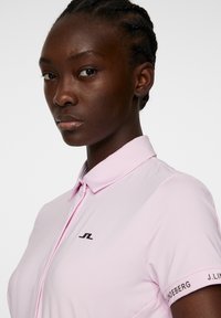 Ljusrosa polo med en subtil lyster, som har en strukturerad krage, svart logga på bröstet och loggdetalj på ärmen.