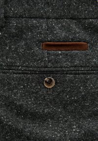 Tela gris oscuro con una superficie texturizada, que presenta un acento de cuero marrón y un botón redondo. Indica un bolsillo cerca de la parte superior.