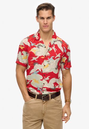 Rød skjorte med korte ærmer, der har et kranemotiv i hvide, blå og gule nuancer, med casual krave og træknapper. Kombination med beige bukser.
