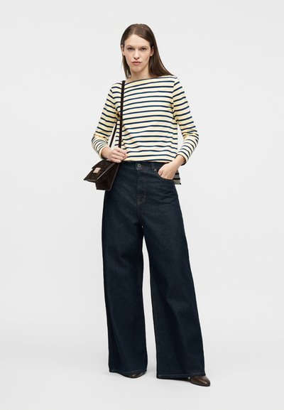 Γυναίκα που φοράει μακρυμάνικο τοπ με κρεμ και ναυτικούς ρίγες, ψηλόμεσα ευρύχωρα σκούρα τζιν, καφέ μπότες και κρατά καφέ τσάντα ώμου.