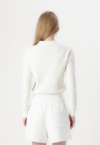 Suéter blanco con textura de canalé, de mangas largas y cuello tipo polo, combinado con pantalones cortos blancos de tiro alto que cuentan con un bolsillo trasero.