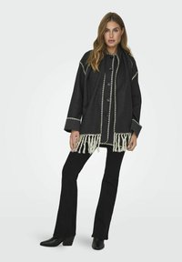 Cappotto in lana nera con ricami bianchi, orlo frangiato e grandi bottoni. Presenta una vestibilità rilassata e maniche larghe. Abbinato a pantaloni neri a zampa.