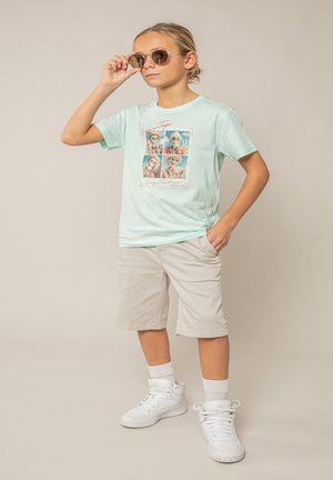 Garçon portant des lunettes de soleil, un T-shirt bleu clair avec des motifs de squelettes, un short beige, des baskets montantes blanches, posant avec la main dans la poche.