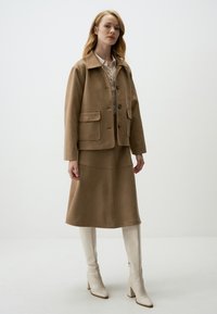 Veste en daim beige avec des boutons en argent et deux poches à l'avant, associée à une jupe évasée assortie. Le mannequin porte des bottes blanches jusqu'au genou.