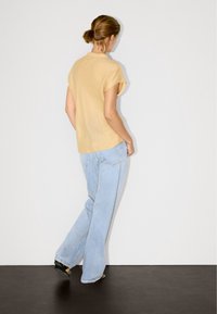 T-shirt di cotone giallo senape a maniche corte, con una vestibilità comoda, abbinata a jeans a vita alta di un blu chiaro, in piedi su un pavimento scuro contro un muro semplice.