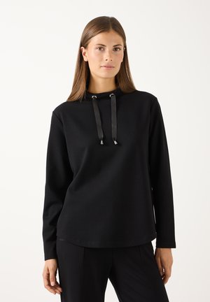 Junge Frau mit glatten braunen Haaren, die einen schwarzen Kapuzenpullover mit langen Ärmeln und Satin-Zügen vor einem schlichten hellen Hintergrund trägt.